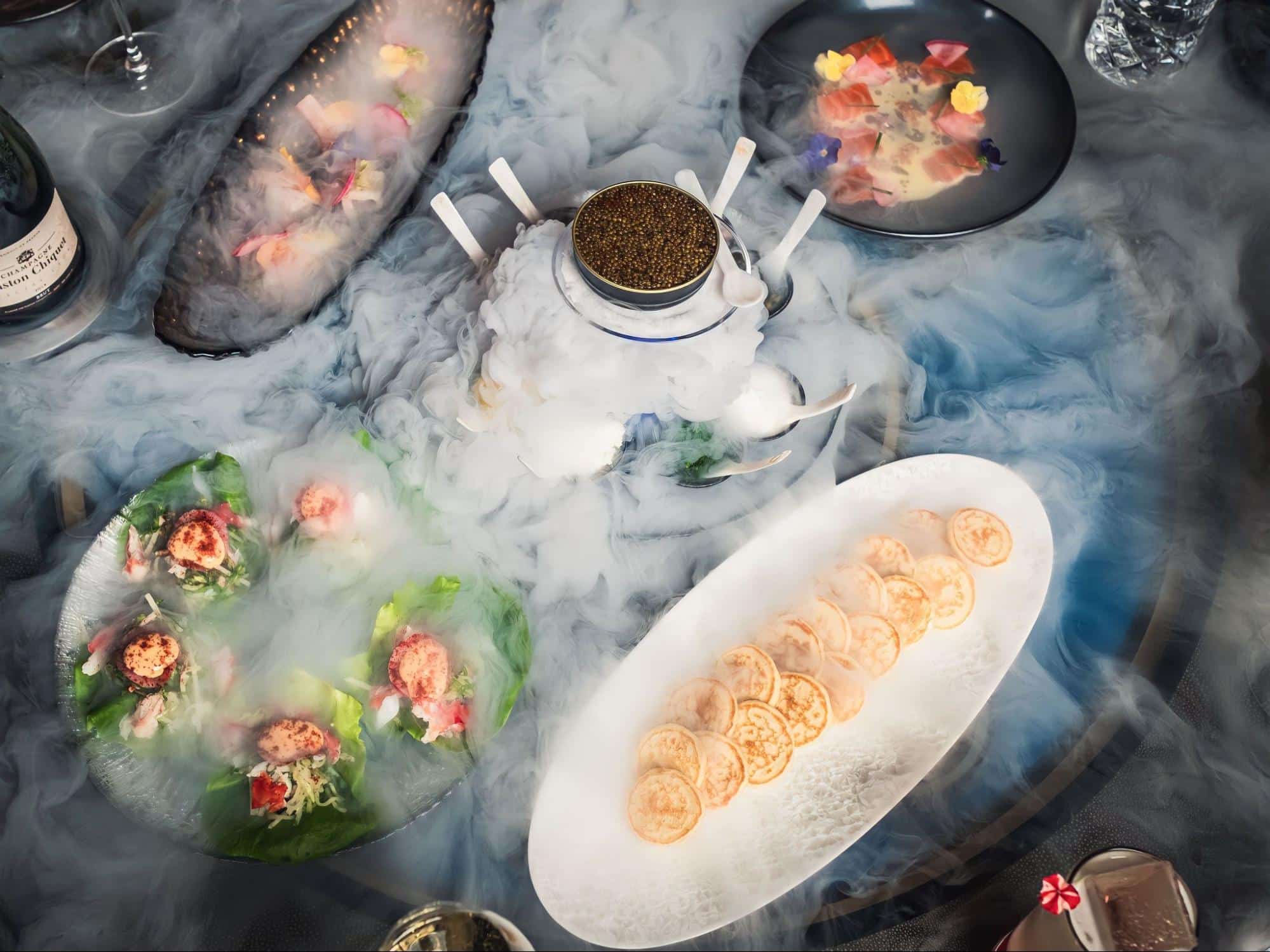 A table full of food from Aqua Seafood & Caviar Restaurant with dry ice surrounding it. 